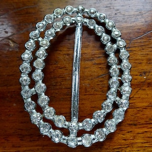 Puede incluir: Hebilla ovalada plateada con dos filas de brillantes redondos y transparentes. Una barra vertical plateada atraviesa el centro de la hebilla. La hebilla está sobre una superficie de madera.