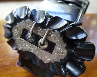 May include: A black belt buckle featuring a scalloped, rounded edge design surrounding a central rectangular silver-toned metal plate. The silver plate is decorated with engraved floral patterns and includes two vertical metal prongs for fastening.