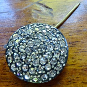 May include: A round, antique-style pin with a dark metal base, encrusted with numerous clear, sparkling rhinestones. A long, gold-colored pin extends from the back. The pin is likely used for decorative purposes.