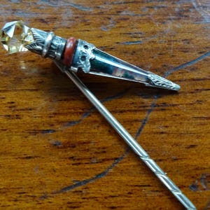 May include: A silver brooch featuring a faceted yellow gemstone. The brooch has a decorative design with a red band and a long, slender pin. The pin is resting on a wooden surface.