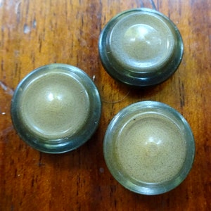 Puede incluir: Tres pomos redondos decorativos con un centro marrón claro y un anillo exterior verde oscuro. Los pomos están sobre una superficie de madera.