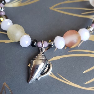 May include: A beaded bracelet featuring a silver teapot charm. The bracelet has white, pink, and yellow beads, with smaller silver and black beads interspersed. The background is a dark gray surface with gold leaf designs.