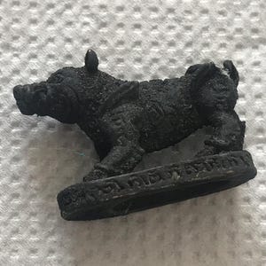 Könnte beinhalten: Eine kleine, dunkel gefärbte Figur eines Wildschweins. Das Wildschwein wird in einer Laufpose dargestellt, mit detaillierter Textur und einem kleinen, kreisförmigen Sockel. Der Sockel hat eine Inschrift. Vor einem weißen, strukturierten Hintergrund.