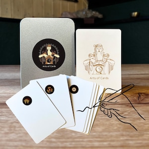 May include: A deck of white cards with gold illustrations and the text "Arts of Cards" is displayed next to a silver tin. The tin has a circular sticker with a woman illustration and the same text. Several cards are fanned out.