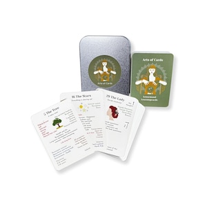 Op de afbeelding: Een set Lenormand-leerkarten met een zilveren blik met een cirkelvormig ontwerp met een figuur. De kaarten zijn wit met tekst en illustraties, waaronder "The Tree" en "The Lady". Het kaartspel is verpakt met een groene kaart.