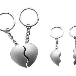 Könnte beinhalten: Zwei silberne Schlüsselanhänger mit herzförmigen Anhängern. Die Anhänger sind in zwei Hälften gebrochen, wobei jede Hälfte an einem separaten Schlüsselanhänger befestigt ist. Die Schlüsselanhänger sind durch eine Kette verbunden.