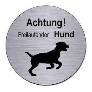Op de afbeelding: Een rond, zilverkleurig bord met de tekst "Achtung! Freilaufender Hund" en een zwart silhouet van een rennende hond.