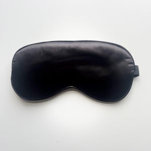 May include: A black satin sleep mask with an elastic strap. The mask is oval and designed to cover the eyes. The sleep mask is a solid black colour and appears to be made of a soft, smooth material.