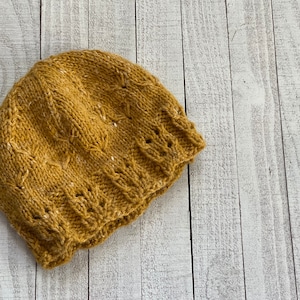 May include: Mustard yellow knit beanie with a textured pattern and small white flecks. The hat features a ribbed brim and a decorative stitch design across the crown. This warm hat is perfect for cold weather.