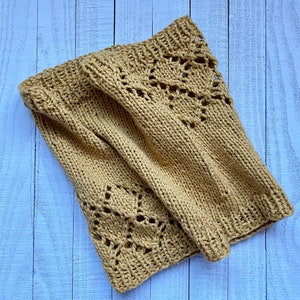 Peut inclure: Une écharpe en tricot beige avec un motif ajouré. L'écharpe est pliée et posée sur une surface en bois bleu clair.