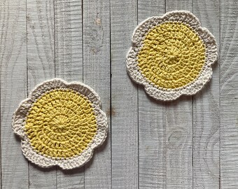 May include: Two crocheted coasters in a flower shape. The coasters are white with a yellow center.