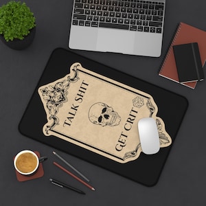 Può includere: Un tappetino per mouse rettangolare nero con una grafica color beige in stile pergamena e bordi neri decorati. Il design include un'illustrazione centrale di un teschio umano, l'icona di un dado a venti facce e il testo "TALK SHIT GET CRIT" in nero.