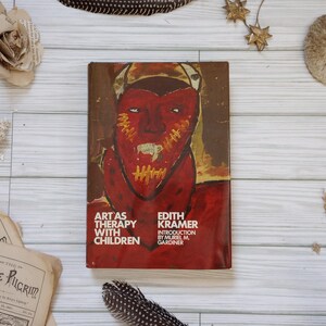 May include: A vintage book titled "Art as Therapy with Children" by Edith Kramer. The cover features a red face with yellow stitching and a brown background. The book is surrounded by decorative elements like paper flowers and feathers.
