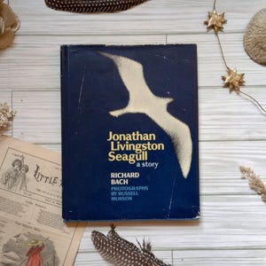 May include: A hardcover book titled Jonathan Livingston Seagull by Richard Bach with photographs by Russell Munson. The cover is dark blue with a large, light-colored silhouette of a seagull in flight and yellow text centered on the front.