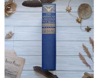 Op de afbeelding: Een vintage hardcover boek getiteld "The American Language" van H.L. Mencken, uitgegeven door Knopf. De omslag van het boek is blauw met gouden letters en een adelaarontwerp.