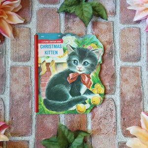 May include: A vintage die-cut children's book titled Christmas Kitten featuring a black and white kitten with a red bow, green pine branches, and yellow ornaments. The book has a red plastic spiral binding and a colorful illustration of a fireplace.