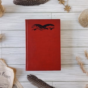 May include: A vintage red book with a black bird illustration on the cover. The book is surrounded by decorative elements, including paper flowers, feathers, and dried plants. The book's title is not visible.