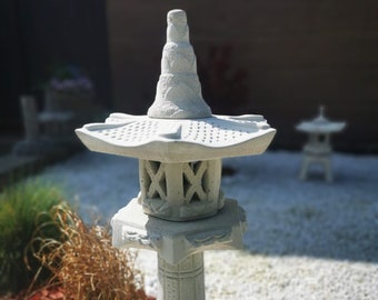 May include: White concrete Japanese lantern with a tiered roof and a decorative base. The lantern is sitting on a bed of white gravel and red mulch.