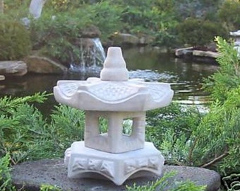 May include: White stone pagoda garden ornament with a traditional design. The pagoda is sitting on a rock near a pond with a small waterfall.