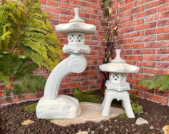 May include: Two white stone Japanese lanterns, one tall and curved, the other shorter and more traditional, set against a brick wall and surrounded by gravel.