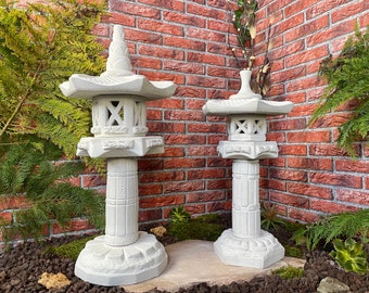 May include: Two white stone Japanese lanterns, one taller than the other, with intricate designs. The lanterns are placed on a bed of gravel and moss in front of a brick wall.