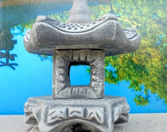 May include: A gray concrete pagoda-style lantern with a square opening in the middle. The lantern is sitting on a textured surface.