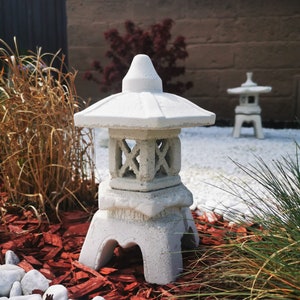 Könnte beinhalten: Zwei weiße japanische Steinlaternen mit einem Pagodendach. Die Laternen stehen auf einem Bett aus roten Holzspänen und weißen Steinen.