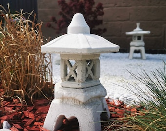 May include: Two white stone Japanese lanterns with a pagoda style roof. The lanterns are sitting on a bed of red wood chips and white rocks.