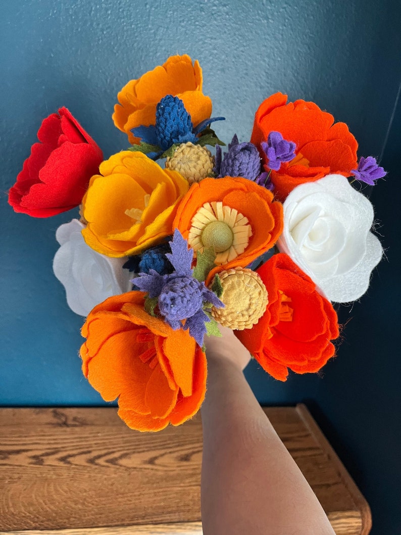 Peut inclure: Un bouquet de fleurs en feutre dans des tons d'orange, de rouge et de blanc. Les fleurs sont dispos&eacute;es en bouquet tenu &agrave; la main et comprennent une vari&eacute;t&eacute; de formes et de tailles de fleurs.