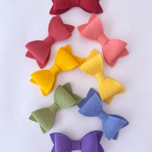 May include: A set of seven felt hair bows in a rainbow color gradient. The bows are arranged in a pyramid shape, with the largest bow at the top and the smallest bow at the bottom. The colors of the bows are red, orange, yellow, green, blue, purple, and pink.