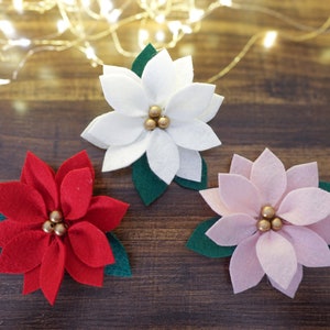 May include: Three felt poinsettia flowers, one red, one white, and one pink, with gold bead centers. The flowers are arranged on a wooden surface.