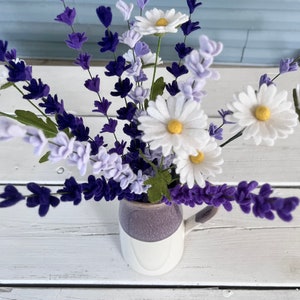 Op de afbeelding: Een boeket van paarse en witte viltbloemen in een witte en paarse keramische mok. De bloemen zijn gerangschikt in een vaas met een witte achtergrond.