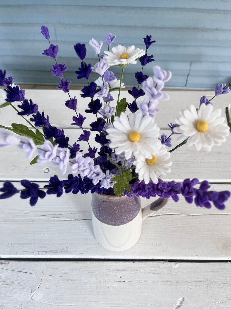 Op de afbeelding: Een boeket van paarse en witte viltbloemen in een witte en paarse keramische mok. De bloemen zijn gerangschikt in een vaas met een witte achtergrond.