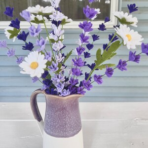 Op de afbeelding: Een boeket van paarse en witte viltbloemen in een paarse en witte gespikkelde keramische kan. De bloemen zijn gerangschikt in een vaas op een witte houten tafel.