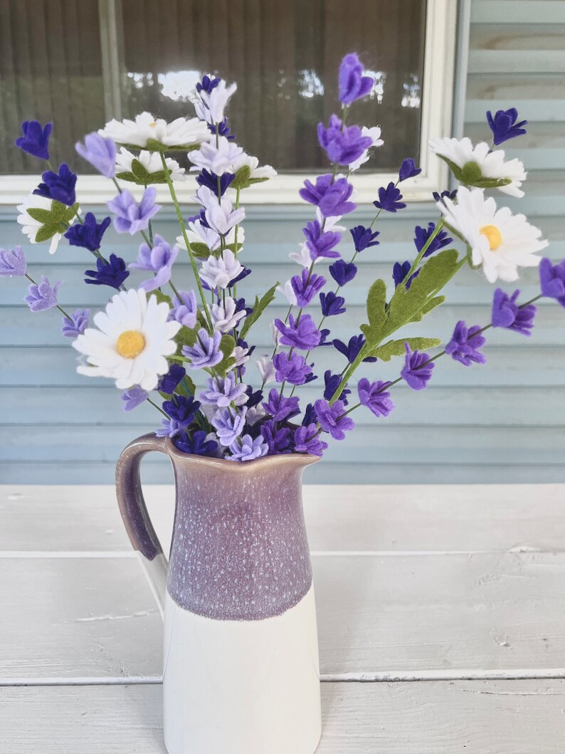 Op de afbeelding: Een boeket van paarse en witte viltbloemen in een paarse en witte gespikkelde keramische kan. De bloemen zijn gerangschikt in een vaas op een witte houten tafel.