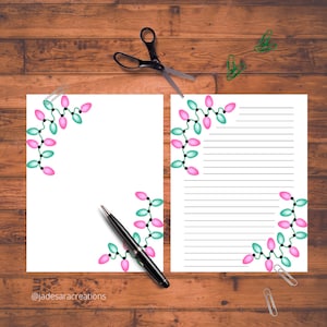 May include: Two white sheets of paper with a Christmas lights design in pink and teal. One sheet is lined, the other is blank. A black pen, scissors, and paper clips are on a wooden surface.