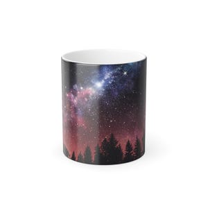 Puede incluir: Una taza de cerámica blanca con un estampado de galaxia negro y morado. El estampado presenta una silueta de un bosque contra un cielo nocturno estrellado.