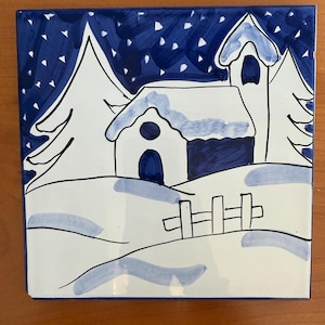 May include: Square ceramic tile with a hand-painted winter scene. The design features a white house and trees against a blue sky with falling snow. A small fence is in the foreground. The tile is approximately 6 inches square.