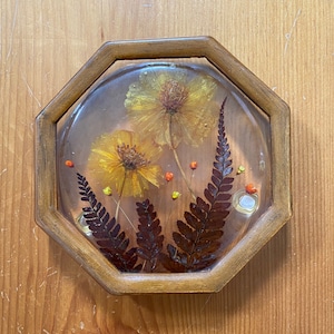 May include: A round, octagonal wooden coaster with a clear resin top. The resin contains pressed dried flowers and ferns in shades of yellow, orange, and brown.