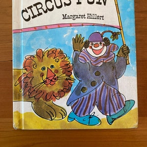 Puede incluir: Una portada de libro infantil vintage con el título "CIRCUS FUN" de Margaret Hillert. La portada presenta una ilustración colorida de un payaso sosteniendo una bandera y un león.