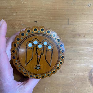 Op de afbeelding: Een ronde houten doos met een geschulpte rand. Het deksel heeft een ingelegd ontwerp van drie figuren met witte cirkels en een ster. De rand heeft zwarte en groene accenten. De doos wordt door een hand vastgehouden.