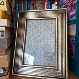 May include: A gold-coloured rectangular picture frame with a patterned interior. The frame is on a wooden surface, with books in the background. The frame has a detailed, textured design.