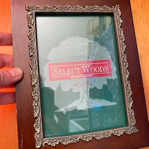 May include: A dark brown wooden picture frame with a gold ornate border. The frame has a green mat and a clear glass front. The frame has a white tree design with the text "SELECT WOODS GENUINE WALNUT 5" x 7" printed on a red label.
