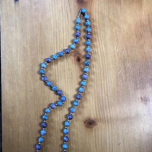 May include: A beaded necklace featuring alternating blue and purple beads with silver-colored spacers. The necklace is displayed on a wooden surface, showcasing its colors and design. The beads have a speckled appearance.