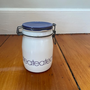 Puede incluir: Un tarro de cerámica blanco con tapa azul y cierre metálico. El tarro tiene la palabra "teateated" impresa en un patrón repetido. El tarro está sobre una superficie de madera.