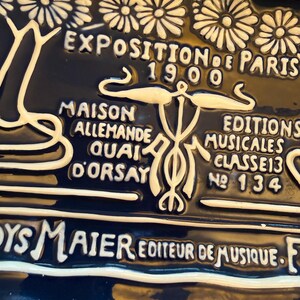 May include: A dark blue ceramic plaque with white raised lettering and floral designs. The text reads "EXPOSITION DE PARIS 1900", "MAISON ALLEMANDE QUAI D'ORSAY", and "EDITIONS MUSICALES CLASSE 13 No 134".