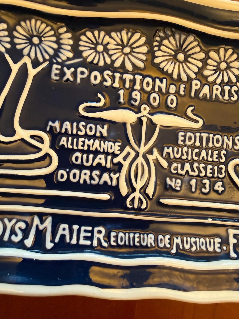 May include: A dark blue ceramic plaque with white raised lettering and floral designs. The text reads "EXPOSITION DE PARIS 1900", "MAISON ALLEMANDE QUAI D'ORSAY", and "EDITIONS MUSICALES CLASSE 13 No 134".