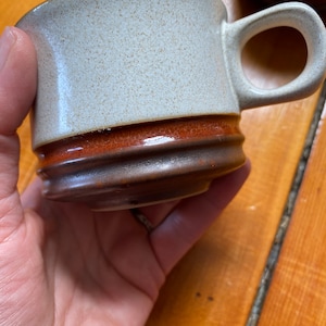 Puede incluir: Una taza de cerámica blanca con un borde marrón y un asa marrón. La taza tiene una textura moteada.