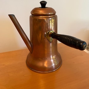May include: A copper coffee pot with a black wooden handle and a black knob on the lid. The pot has a spout and a rounded base.