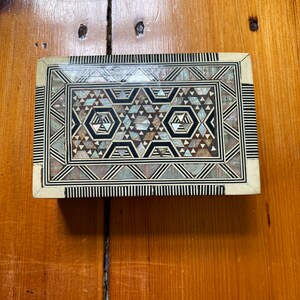 Op de afbeelding: Een kleine houten doos met een geometrisch patroon ingelegd met parelmoer. De doos is gemaakt van licht hout met zwarte en witte accenten.